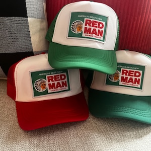 May include: Three trucker hats with white mesh backs and brims in red and green. Each hat features a white square patch with the text "RED MAN CHEWING TOBACCO" and an image of a Native American.