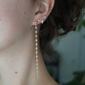 May include: A close-up of an ear adorned with multiple gold earrings. The earrings include a long, dangling chain earring, a small hoop earring with a clear stone, a star-shaped stud, a small stud, and a rectangular bar earring.