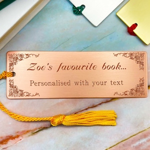 Könnte beinhalten: Kupferfarbener Metall-Lesezeichen mit dekorativen Eckdesigns und dem Text "Zoe's favourite book... Personalised with your text." Ein gelber Quastenanhänger ist befestigt.