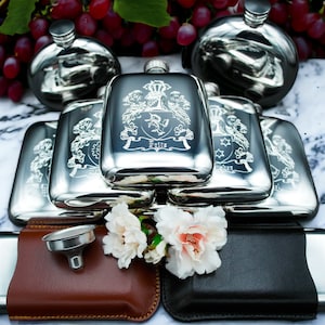 Peut inclure: Une pile de flacons à liqueur en argent avec des motifs gravés, dont un blason avec le nom "Eytle" et un entonnoir. Les flacons sont sur une surface blanche avec un étui en cuir marron et un étui en cuir noir.