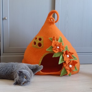 Puede incluir: Cama para gatos de fieltro naranja con forma de casa con hojas de fieltro verde y flores de fieltro naranja. Un gato gris está acostado frente a la cama.
