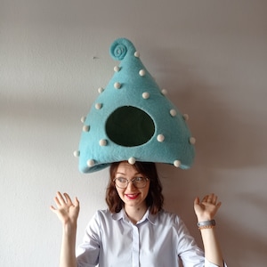 Può includere: Una donna indossa un cappello per gatti in feltro blu chiaro con pompon bianchi. Il cappello ha una cima arrotondata e una punta arricciata. La donna sorride e ha le mani alzate.