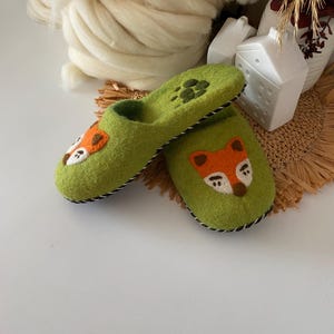 May include: Handmade green felt slippers with fox faces and paw prints. The slippers have orange, white, and black details, and a black and white stitched trim. These cozy slippers are perfect for indoor wear.