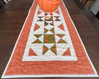 May include: A quilted table runner with a fall theme. The runner is made of orange, white, and green fabric and features a star pattern with pumpkins. A ceramic pumpkin sits on top of the runner.