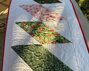 May include: A white table runner with a red border and a quilted design. The runner features a series of green, red, and pink diamond-shaped patches with various Christmas-themed patterns.