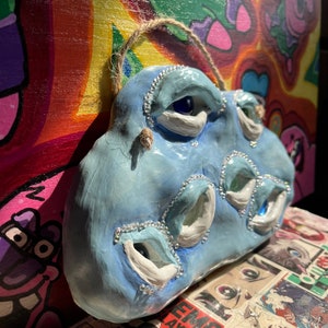 Könnte beinhalten: Eine hellblaue, abstrakte Keramikskulptur mit mehreren augenförmigen Öffnungen. Die Skulptur ist mit kleinen, klaren Strasssteinen um die Augenöffnungen und einer Schnurschlaufe zum Aufhängen verziert. Der Hintergrund zeigt farbenfrohe, psychedelische Kunst.