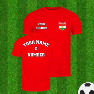 Puede incluir: Dos camisetas rojas con texto blanco sobre un fondo verde. La parte delantera de la camiseta tiene el texto "YOUR NUMBER" y un escudo con la palabra "KURDISTAN" y una bandera. La parte trasera de la camiseta tiene el texto "YOUR NAME & NUMBER". Hay un bal&oacute;n de f&uacute;tbol en la esquina.