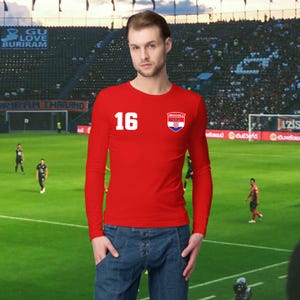 May include: A red long-sleeve shirt with the number 16 in white on the left chest. A Croatian flag emblem is on the right chest. The shirt is paired with blue denim jeans. The background shows a soccer field and stadium.