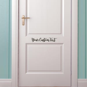 May include: A white door with a gold-colored handle and the text "Your Custom Text" in black script. The door is set against a light blue wall.