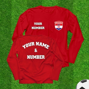 May include: Two red long-sleeve shirts with white text and a Croatian flag emblem. One shirt displays "YOUR NUMBER" and the other says "YOUR NAME & NUMBER". The shirts are on a green background, suggesting a sports theme.