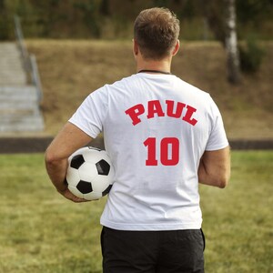 Puede incluir: Una camiseta blanca con el nombre "PAUL" y el n&uacute;mero "10" en letras rojas en la espalda. La persona sostiene un bal&oacute;n de f&uacute;tbol blanco y negro en un campo de hierba.