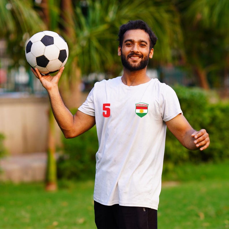 Puede incluir: Un bal&oacute;n de f&uacute;tbol con paneles en blanco y negro se sostiene en el aire. La persona lleva una camiseta blanca con el n&uacute;mero 5 en rojo y un parche con la bandera de Kurdist&aacute;n.