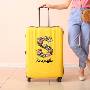 Puede incluir: Una maleta rígida de color amarillo brillante con una letra S grande decorada con un patrón floral colorido. El nombre Samantha está impreso en letra cursiva negra debajo de la letra. La maleta tiene un asa retráctil y ruedas giratorias.