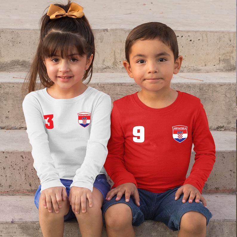 May include: Two children wearing long-sleeved shirts. One child is wearing a white shirt with the number 3 and a Croatian emblem. The other child is wearing a red shirt with the number 9 and a Croatian emblem. Both shirts have the word "Hrvatska" on them.