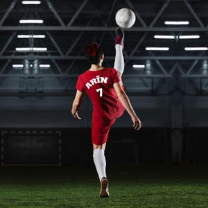 Puede incluir: Una persona con un uniforme de f&uacute;tbol rojo pateando un bal&oacute;n en el aire. El uniforme tiene el nombre "Arin" y el n&uacute;mero "7" en la espalda. La persona lleva calcetines blancos y zapatos negros y rojos. El fondo es un campo de f&uacute;tbol.