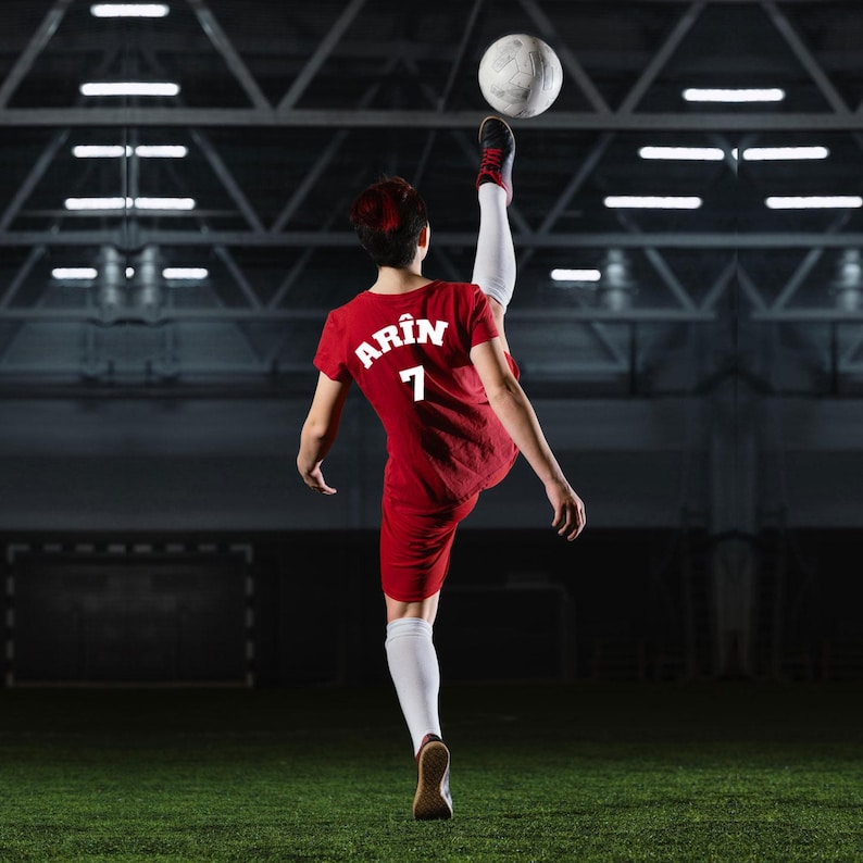 Puede incluir: Una persona con un uniforme de f&uacute;tbol rojo pateando un bal&oacute;n en el aire. El uniforme tiene el nombre "Arin" y el n&uacute;mero "7" en la espalda. La persona lleva calcetines blancos y zapatos negros y rojos. El fondo es un campo de f&uacute;tbol.