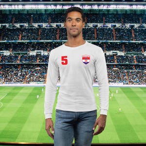 May include: White long-sleeve shirt with the number 5 in red on the left chest. A Croatian crest is on the right chest. The shirt is worn with blue jeans. The background is a soccer stadium.