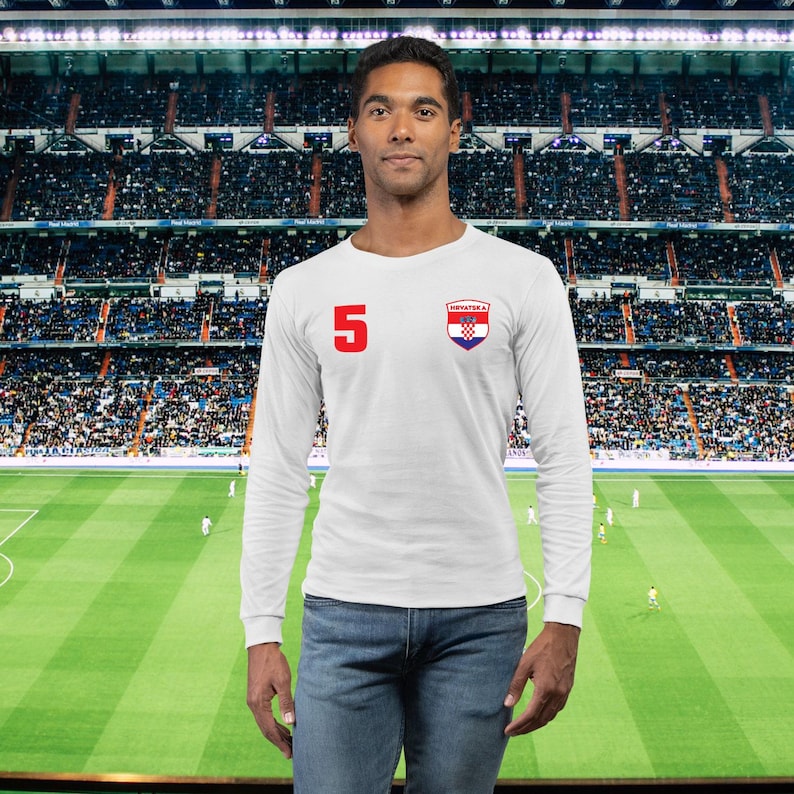 May include: White long-sleeve shirt with the number 5 in red on the left chest. A Croatian crest is on the right chest. The shirt is worn with blue jeans. The background is a soccer stadium.