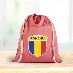 May include: Red drawstring backpack with the word "ROMÂNIA" above a shield displaying the Romanian flag. The flag features vertical stripes of blue, yellow, and red. The bag is sitting on a wooden surface.