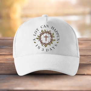 May include: White baseball cap with a curved peak. The cap displays a circular design with a cross, a crown of thorns, and the text "A LOT CAN HAPPEN IN 3 DAYS". The design is in brown and green.