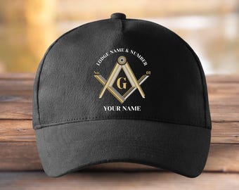 May include: Black baseball cap with a Masonic emblem. The emblem features a compass and square design with the letter "G" in the center, and the text "Lodge Name & Number" and "Your Name". The cap is on a wooden surface.