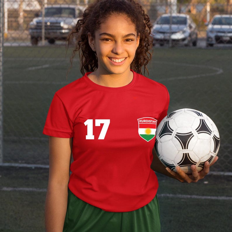 Puede incluir: Una camiseta de f&uacute;tbol roja con el n&uacute;mero 17 en blanco, un parche con la bandera de Kurdist&aacute;n y pantalones cortos verdes. Un bal&oacute;n de f&uacute;tbol se sostiene frente a la camiseta. El fondo muestra un campo de f&uacute;tbol y coches aparcados.
