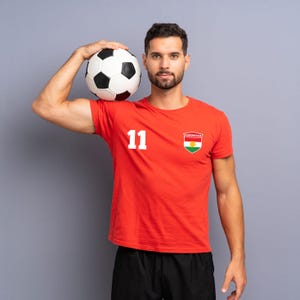 Puede incluir: Una persona sostiene un bal&oacute;n de f&uacute;tbol blanco y negro y lleva una camiseta roja con el n&uacute;mero 11 y un parche con la bandera de Kurdist&aacute;n. La persona tambi&eacute;n lleva pantalones cortos negros. Fondo gris.