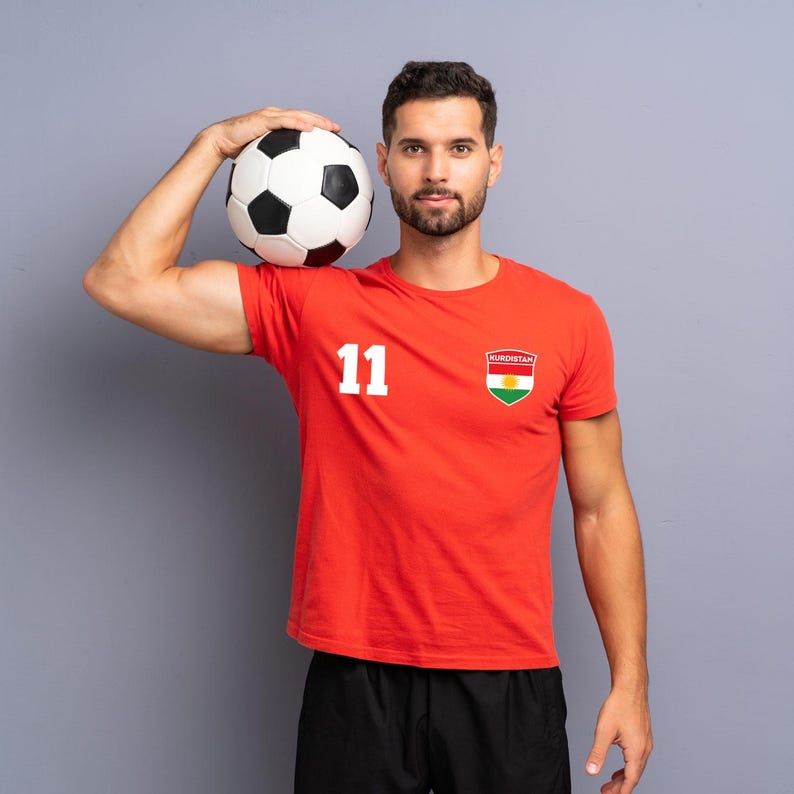 Puede incluir: Una persona sostiene un bal&oacute;n de f&uacute;tbol blanco y negro y lleva una camiseta roja con el n&uacute;mero 11 y un parche con la bandera de Kurdist&aacute;n. La persona tambi&eacute;n lleva pantalones cortos negros. Fondo gris.