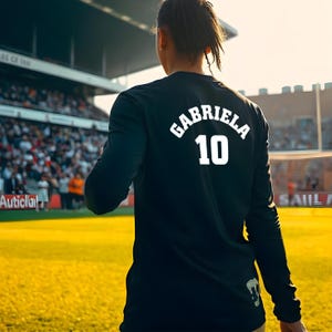 May include: A black long-sleeve shirt with the name "GABRIELA" and the number "10" printed in white on the back. The shirt is worn by a person on a soccer field, with a stadium and crowd in the background.