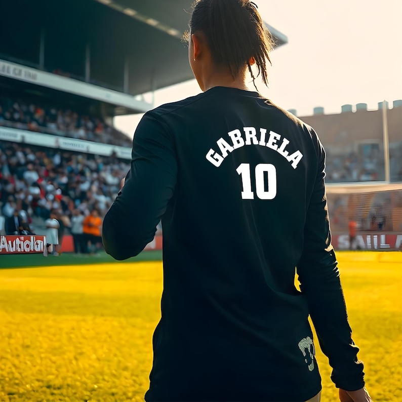 May include: A black long-sleeve shirt with the name "GABRIELA" and the number "10" printed in white on the back. The shirt is worn by a person on a soccer field, with a stadium and crowd in the background.
