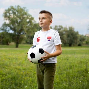 K&ouml;nnte beinhalten: Ein junger Junge tr&auml;gt ein wei&szlig;es Fu&szlig;balltrikot mit der Nummer 5 und der kroatischen Flagge auf der Brust und h&auml;lt einen Fu&szlig;ball vor sich. Er steht auf einem grasbewachsenen Feld.