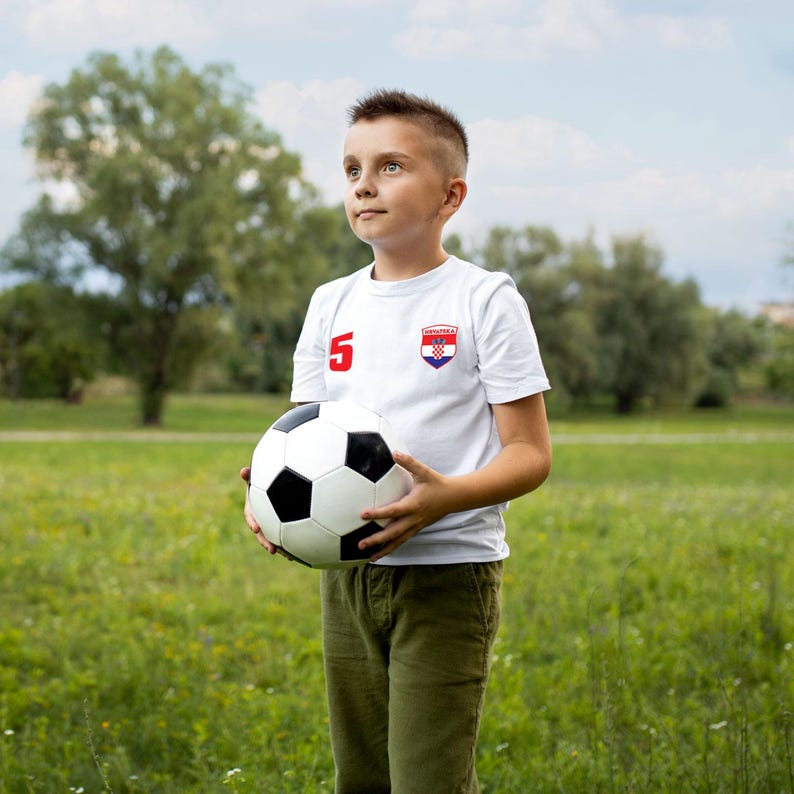 K&ouml;nnte beinhalten: Ein junger Junge tr&auml;gt ein wei&szlig;es Fu&szlig;balltrikot mit der Nummer 5 und der kroatischen Flagge auf der Brust und h&auml;lt einen Fu&szlig;ball vor sich. Er steht auf einem grasbewachsenen Feld.