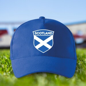 Könnte beinhalten: Eine königsblaue Baseballkappe mit dem Emblem der schottischen Flagge. Das Emblem zeigt ein weißes Andreaskreuz auf einem blauen Schild, darüber steht das Wort "SCOTLAND". Der Hintergrund ist verschwommen, mit grünem Gras.