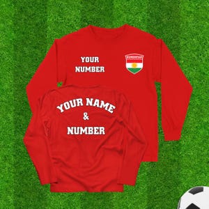 Może przedstawiać: Czerwona koszulka z długim rękawem z napisem "YOUR NUMBER" z przodu i "YOUR NAME & NUMBER" z tyłu. Z przodu znajduje się również emblemat flagi Kurdystanu. Koszulka jest pokazana na zielonym tle.