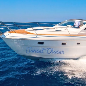 May include: A white motorboat with a wooden deck is sailing on blue water under a clear sky. The boat has the text "Sunset Chaser" in blue script on its side. The boat is moving fast, creating a wake.
