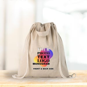 Op de afbeelding: Een beige trekkoordtas met een kleurrijk logo met de tekst "YOUR PHOTO TEXT LOGO MONOGRAM" en "FRONT & BACK SIDE". De tas heeft een trekkoordsluiting en een klein handvat aan de bovenkant. De tas staat op een houten oppervlak.