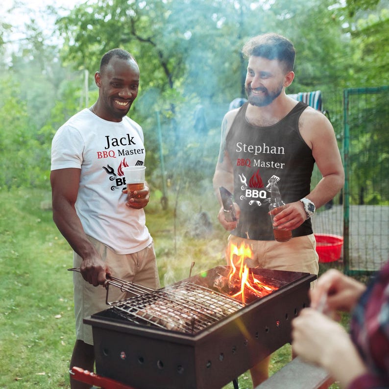 May include: Two people at a barbecue, one wearing a white t-shirt with "Jack BBQ Master" printed on it, and the other wearing a black tank top with "Stephan BBQ Master". A grill with flames and food is in the foreground.