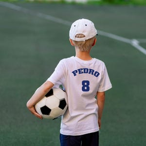 May include: A young person wearing a white t-shirt with the name "PEDRO" and the number "8" on the back, holding a soccer ball. The person is wearing a white baseball cap and is standing on a green field.