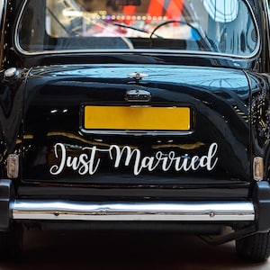 Könnte beinhalten: Rückseite eines schwarzen Taxis mit dem Schriftzug "Just Married" in weißer Schrift. Das Auto hat ein gelbes Nummernschild und eine Chromstoßstange. Die Aufnahme erfolgt aus niedriger Perspektive.