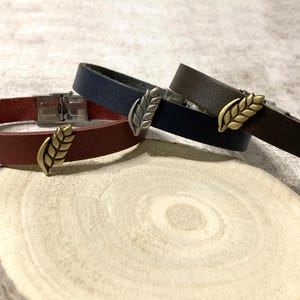 Puede incluir: Tres pulseras de cuero en burdeos, azul marino y marrón, cada una con un adorno de hoja metálica. Las pulseras se exhiben sobre una rodaja de madera, mostrando los diferentes colores y diseños. Las pulseras tienen un diseño sencillo y elegante.