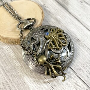 Könnte beinhalten: Eine silberne Taschenuhr mit Kette, die mit einem komplizierten Design eines Kraken mit einem blauen Edelsteinauge verziert ist. Das Zifferblatt der Uhr ist mit römischen Ziffern sichtbar. Der Krake ist von einem dekorativen Rand umgeben.