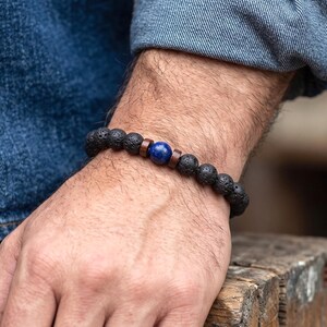Peut inclure: Un bracelet composé de perles rondes en pierre de lave noire poreuse, agrémenté de deux petites entretoises en bois brun et d'une perle unique, lisse et ronde, en lapis-lazuli bleu profond, placée au centre du cordon élastique.