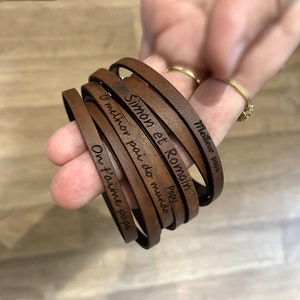Könnte beinhalten: Ein Set aus braunen Lederarmbändern mit gravierten Botschaften, darunter "Simon et Romain", "Poppy", "O melhor pai do mundo" und "On t'aime papa".