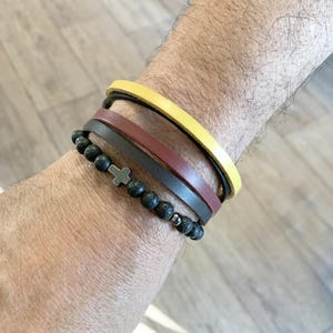Può includere: Una collezione di braccialetti su un polso. I braccialetti includono una fascia in pelle gialla, una fascia in pelle bordeaux, una fascia in pelle grigio scuro e un braccialetto di perline nere con un ciondolo a croce argentata.