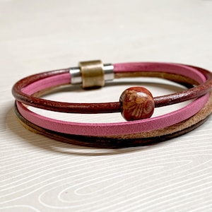 Op de afbeelding: Een leren armband met meerdere strengen en een magnetische sluiting. De armband heeft drie strengen in bruin-, roze- en beigetinten. Een houten kraal met een decoratief patroon is gecentreerd op de armband.