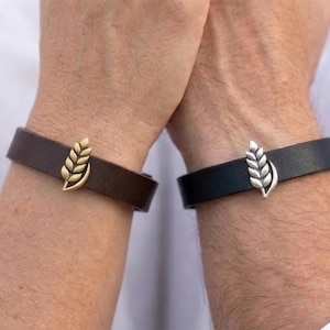 Pode incluir: Duas pulseiras de couro, uma castanho-escura com um pendente de espiga de trigo em tom dourado e uma preta com um pendente de espiga de trigo em tom prateado. Cada pulseira apresenta uma tira de couro lisa e um emblema metálico central.