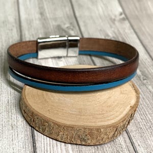 Könnte beinhalten: Ein braunes Lederarmband mit blauem Akzentband und einem silbernen Magnetverschluss. Das Armband wird auf einem kleinen, runden Holzuntersetzer präsentiert, der ein minimalistisches Design zeigt. Ein stilvolles Accessoire.