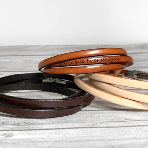 Peut inclure: Trois bracelets enroulants en cuir dans les tons marron et beige. Le bracelet supérieur est marron moyen avec l'inscription gravée "parce que tu es unique". Les deux autres bracelets sont marron foncé et beige clair. Chaque bracelet a un fermoir argenté.
