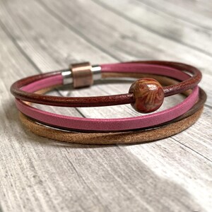 Może przedstawiać: Wielopasmowa skórzana bransoletka z magnetycznym zapięciem i ozdobnym drewnianym koralikiem. Bransoletka ma trzy pasma w odcieniach brązu i różu. Drewniany koralik jest w jasnobrązowym kolorze z wzorem słojów drewna.