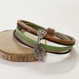 May include: A multi-strand bracelet with brown, green, and tan leather cords. A silver-colored charm in the shape of a leaf and a heart hangs from the bracelet. The bracelet is resting on a small, round wooden slice.